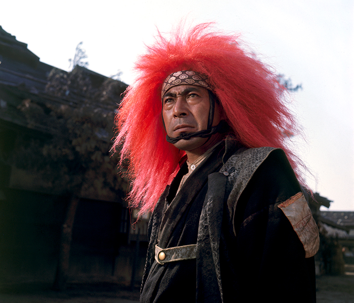 Still of a person staring intensely and wearing a red lion’s mane and a patterned headband; a village and trees are in the background. 