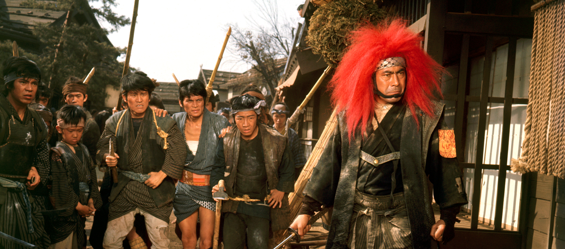 Still of a group of people of ranging ages carrying staffs as weapons; they are led by the person in front of them wearing a red lion’s main and patterned headband, holding a sword.
