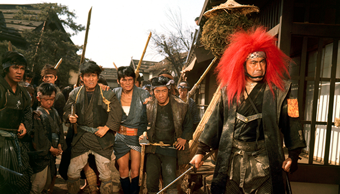 Still of a group of people of ranging ages carrying staffs as weapons; they are led by the person in front of them wearing a red lion’s main and patterned headband, holding a sword.