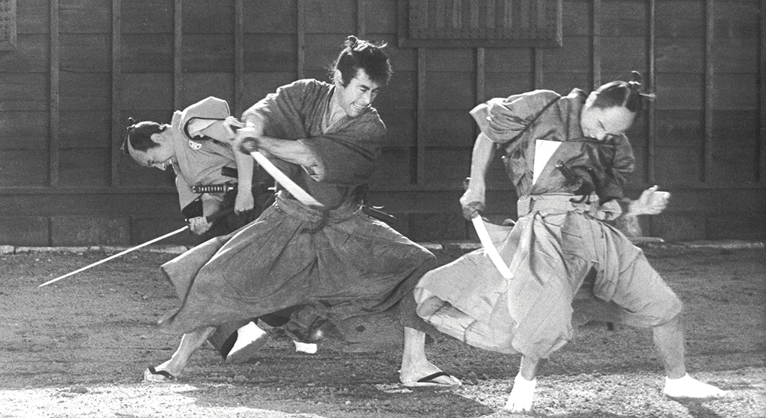 Black-and-white still of a fight scene; the person in the middle is lunging with their sword toward the person in front of them; the third person in the back is facing the other direction, their sword and head pointed toward the ground.