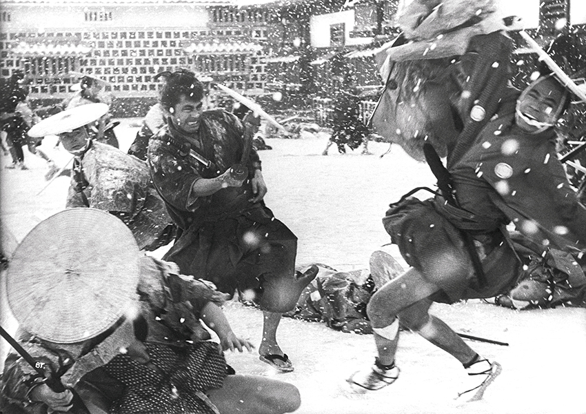Black-and-white still of a fight scene in the snow; two people in front are falling to the ground while being chased by people with swords.