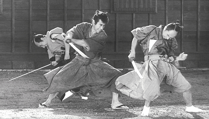 Black-and-white still of a fight scene; the person in the middle is lunging with their sword toward the person in front of them; the third person in the back is facing the other direction, their sword and head pointed toward the ground.
