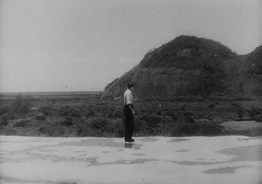 Black-and-white still of a person wearing a white shirt and black pants; they are standing outside looking towards the rocky hill and field in the background.