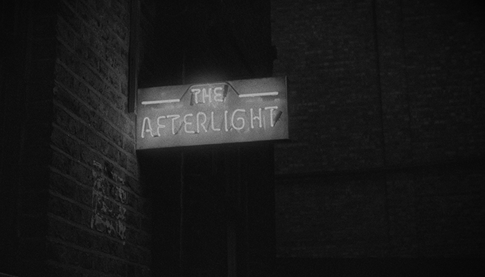 Black-and-white still of a neon-lit sign that says The Afterlight; it is protruding from a brick wall.