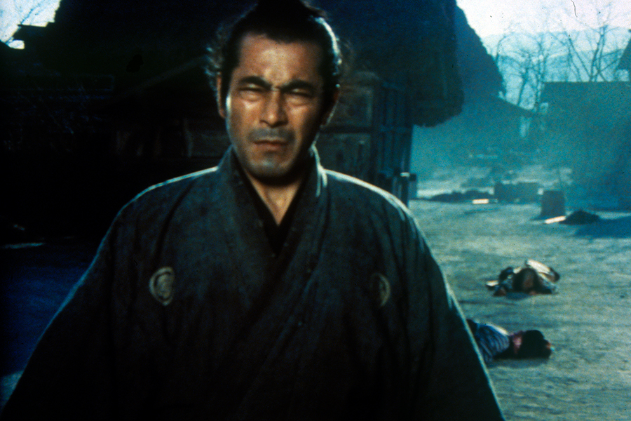 Still of a person wearing a kimono and squinting in the sun; there are bodies on the ground behind them. 