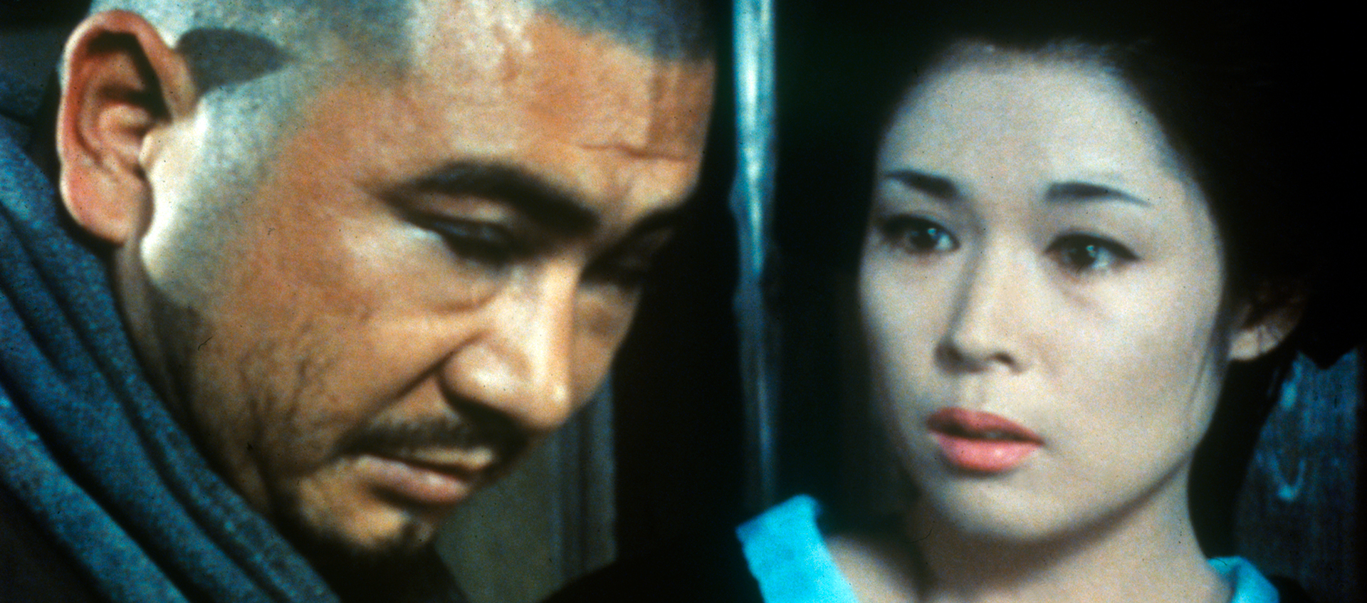 Still of a person with dark hair, pink lipstick, and a blue-lined kimono looking concernedly at a person with a shaved head and mustache whose head is pointed down and eyes are closed. 