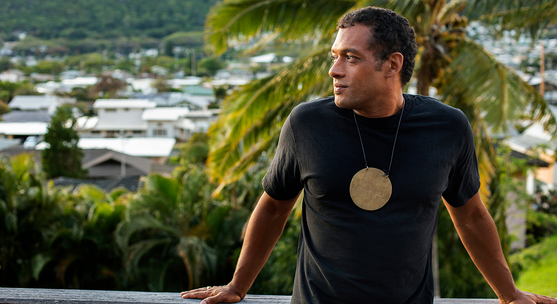 Makaya McCraven, who has dark hair and is wearing a large golden circle pendant over a black shirt, stands, looking to the left, in front of tropical greenery and houses in the background. 