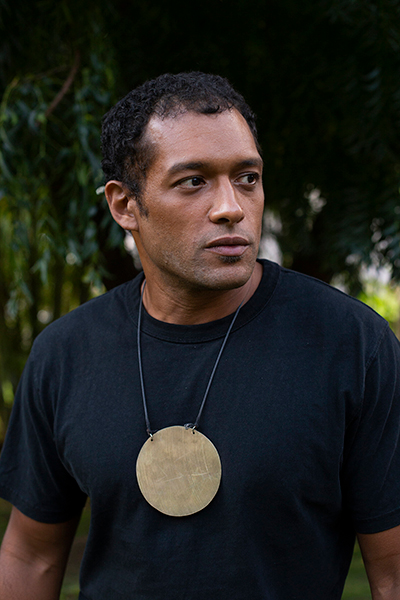 Makaya McCraven, who has dark hair and is wearing a large golden circle pendant over a black shirt, stands, looking to the right, in front of tropical foliage. 