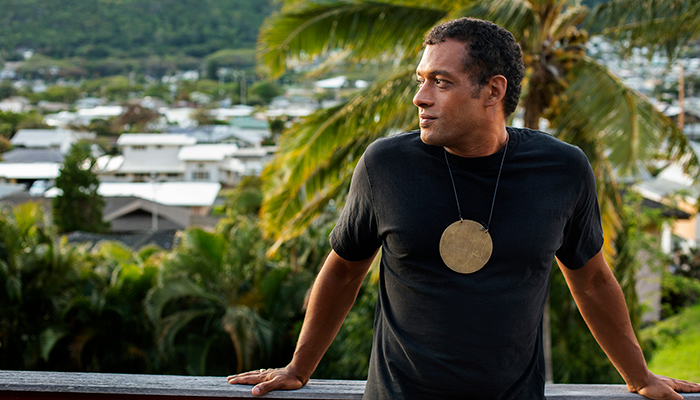 Makaya McCraven, who has dark hair and is wearing a large golden circle pendant over a black shirt, stands, looking to the left, in front of tropical greenery and houses in the background. 