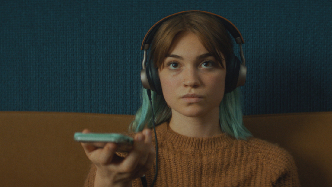 A young white woman with light brown hair with green tips is presented from shoulders up, sitting on a beige couch. She's wearing headphones and holding a green smartphone. She's looking up at something behind the camera.