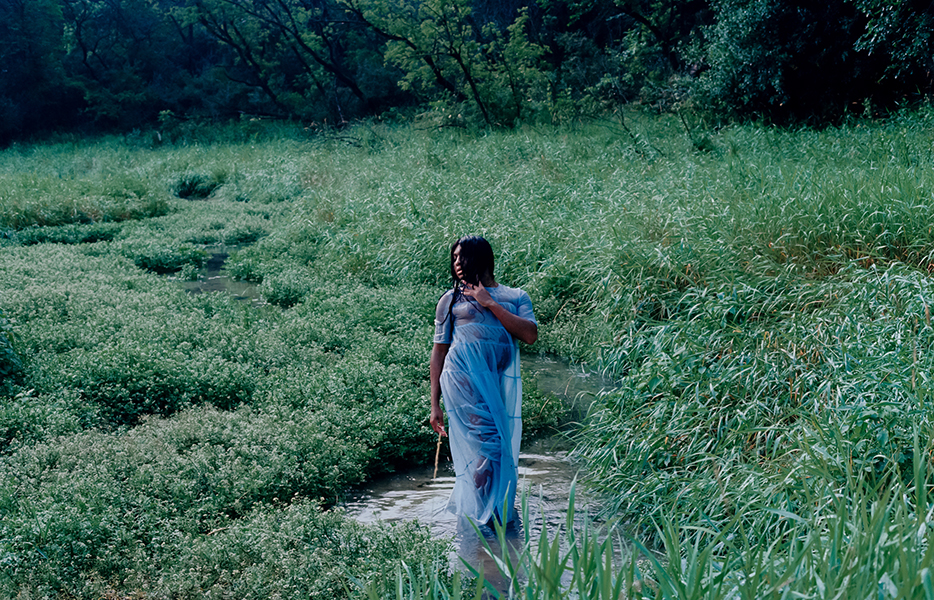 A still image from artist Keioui Keijaun Thomas's video Comme Hell or High Femmes: Act 2. The Last Trans Femmes on Earth: Dripping Doll Energy. In the foreground, Thomas stands in a pool of water at the edge of a stream surrounded by green grasses. She has brown skin and long brown hair, and she is wearing a long, transparent, wet blueish dress and is dangling a stick from her right-hand fingers. In the background is a line of leafy green trees.