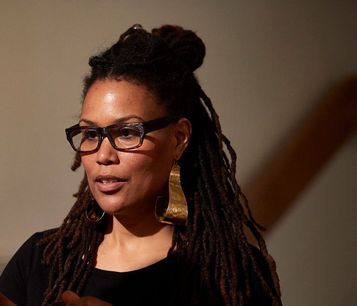 Photo of artist Jasmine Murrell, who has brown skin and long, dark brown hair, half of which is pull back in a bun. She is wearing a black shirt, black glasses, and gold pendant earrings. 