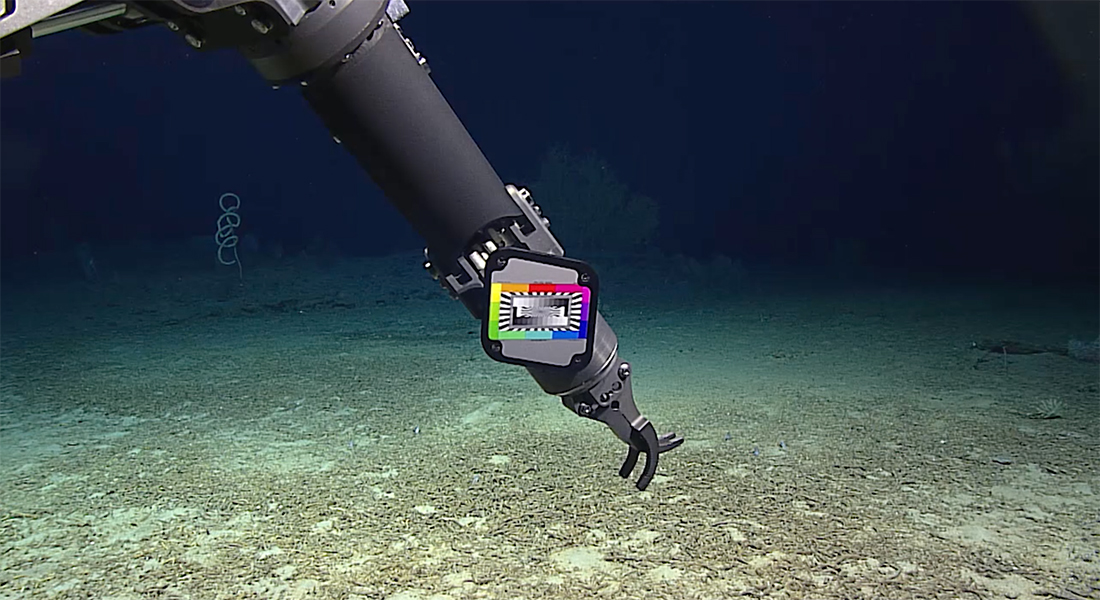 Still of a robotic arm with a claw extending towards the blue, green, and brown ocean floor with deep blue, opaque water in the background.