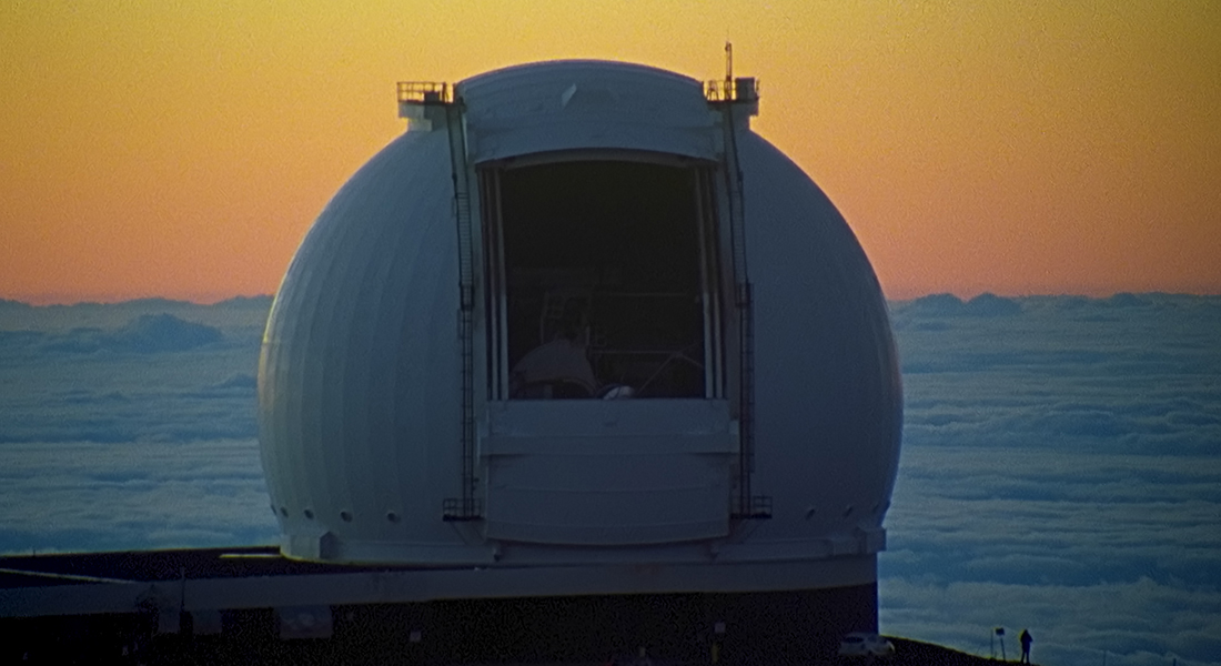 Still of large, white dome observatory on a platform above the pillowy, light blue clouds in the background below the orange-yellow sky. To the bottom right of the dome and platform is a silhouette of a person who appears minuscule in comparison.