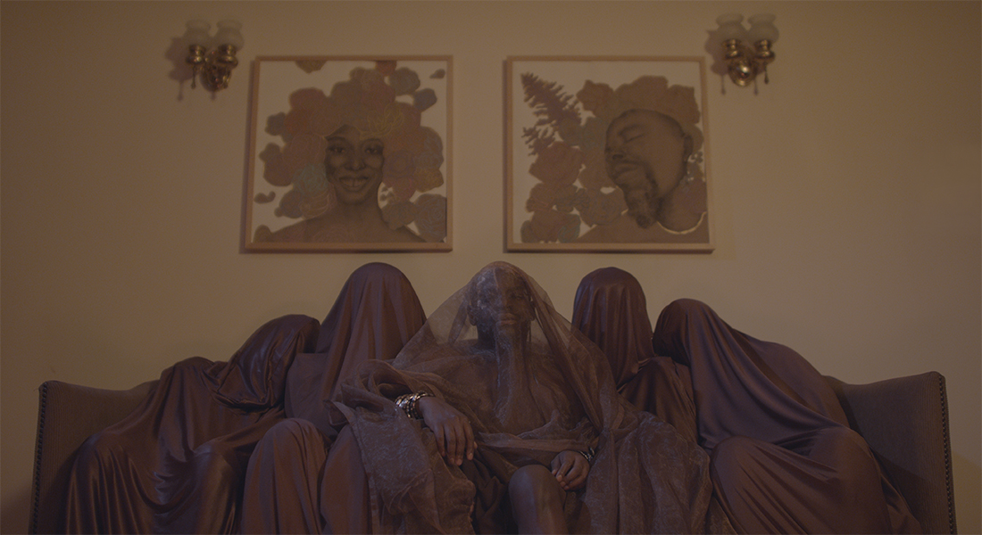 Five figures, each of whom are draped in brown fabric, sit on a brown couch. The person in the center is draped in transparent brown fabric, revealing their face and dark brown skin. Their hands, which are uncovered, rest on their legs, and there are many shiny bracelets on both wrists. Above the figures, on the wall behind them, are two portraits of Black people, both of whom have flowers and leaves surrounding their heads like hair. 