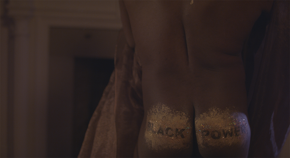 The bare backside of a Black person, who is holding transparent brown fabric, with the words “BLACK POWER” written in black against a sparkly gold background painted on the person’s bottom.