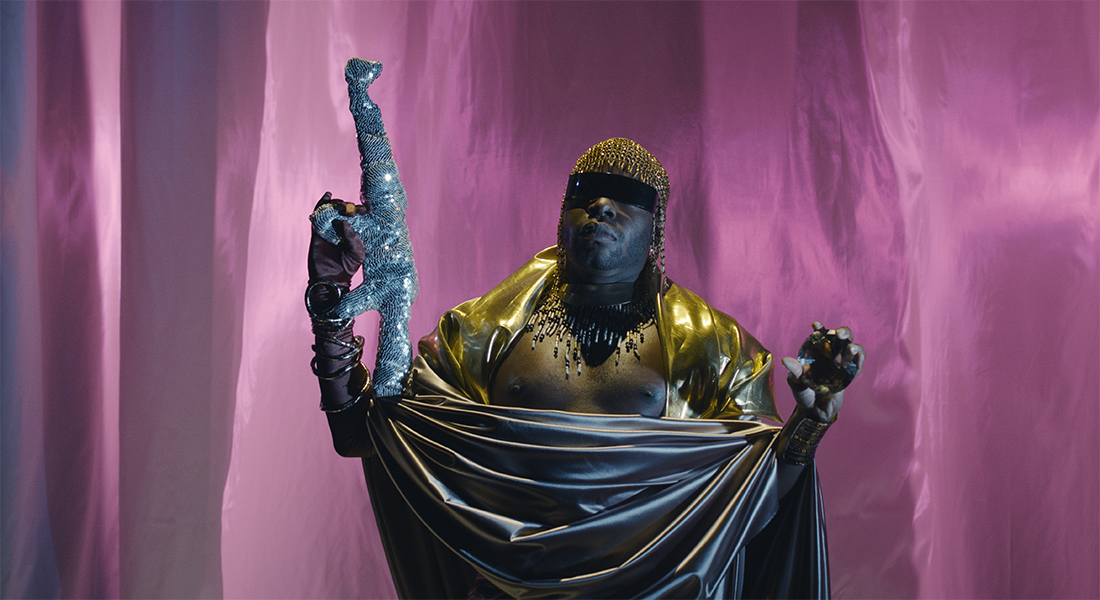 jaamil olawale kosoko standing against a background of pink, shimmery fabric. They have a black blindfold covering their eyes and are wearing a gold chainmail headpiece; a wide, gold metal necklace with light-and-dark beads hanging from it; a brown satin glove and gold, coiled arm band on one arm and a golden cuff on the other, and goldish brown fabric draped over their shoulders and stomach. They are holding a silver, sequenced object that resembles a gun in one hand and a golden-brown jewel in the other. 