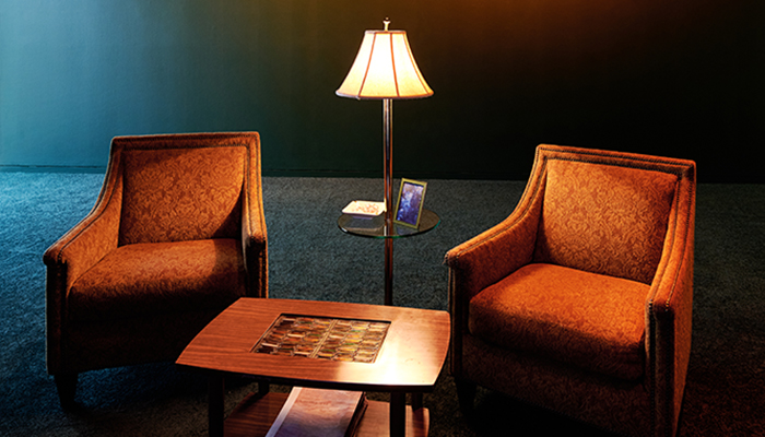 Gallery space with green walls and carpet, featuring two orangish red armchairs with floral pattering and two rows of gold studs embroidered along the edges; a coffee table; and a tall, lit lamp with a picture frame and bowl of mints resting on the small glass table attached to the lamp pole.