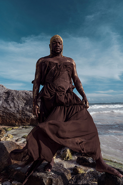 jaamil olawale kosoko from their 2022 book Black Body Amnesia: Poems and Other Acts, featuring the artist standing against a rocky ocean inlet. They are wearing a gold chainmail headpiece, gold body paint on their face and torso, and a flowing brown gown.