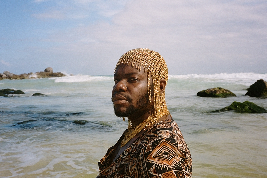 Artist jaamil olawale kosoko from their video project Syllabus for Black Love. The artist wears a gold, chainmail headpiece and a black-and-gold patterned robe. Behind them in the background is a rocky ocean and blue sky with light clouds.