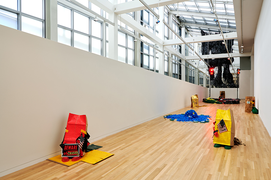 Gallery space with white walls and wooden floors, featuring an installation made of red and yellow paper bags, dead leaves, American flags, trash bags, red lights, and other miscellaneous objects.