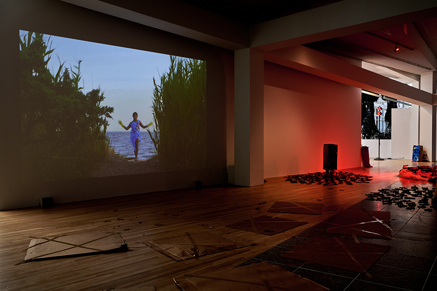 Gallery space that is dimly lit with red light. The projection on the wall features Keioui Keijaun Thomas standing on a sandy beach, in between tall grass and trees. The ocean and blue sky are behind her. Square pieces of cardboard are taped to the wooden gallery floor in a seating arrangement.