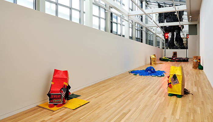 Gallery space with white walls and wooden floors, featuring an installation made of red and yellow paper bags, dead leaves, American flags, trash bags, red lights, and other miscellaneous objects. 