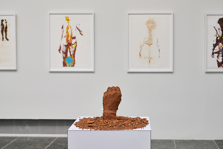 Sculpture made of dirt in the shape of a fist on a white pedestal in a gallery space. The backdrop is a white wall with drawings of female figures in white frames.