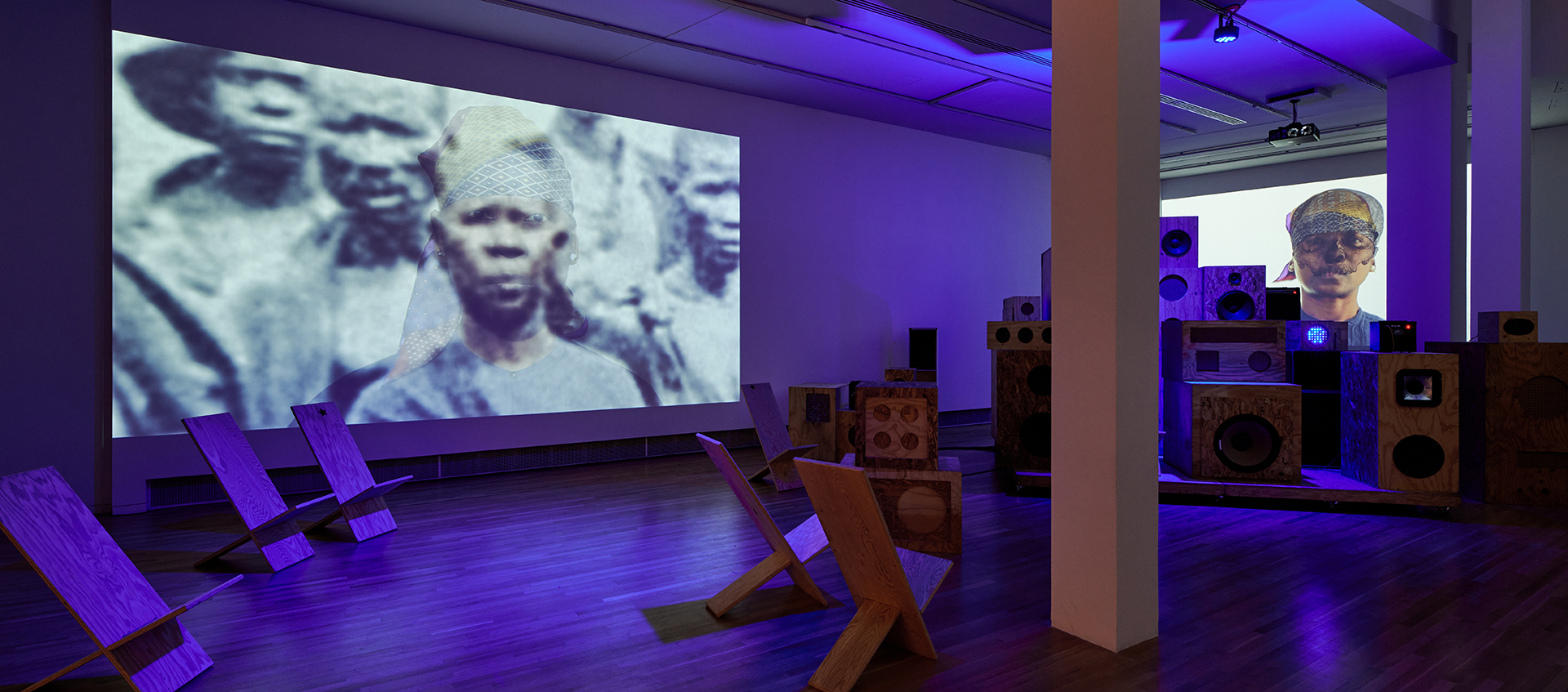 Gallery space lit with purple light. Multisized wooden speakers are stacked on top of each other in the back right corner. Behind them is a projection of nora chipaumire with a black scull covering her face and pattered headscarf. There are wooden chairs scattered throughout the wooden gallery floors to the left of the speaker system. Above them is a larger projection featuring a black-and-white photo of a group of African people; nora’s face photoshopped in color over someone else’s.