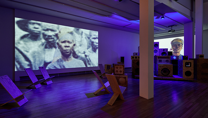 Gallery space lit with purple light. Multisized wooden speakers are stacked on top of each other in the back right corner. Behind them is a projection of nora chipaumire with a black scull covering her face and pattered headscarf. There are wooden chairs scattered throughout the wooden gallery floors to the left of the speaker system. Above them is a larger projection featuring a black-and-white photo of a group of African people; nora’s face photoshopped in color over someone else’s.