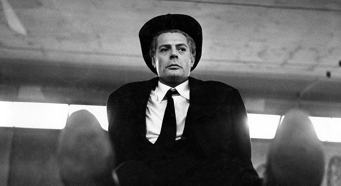 Black-and-white still of Guido (Marcello Mastroianni) sitting with legs outstretched, wearing a black suit, tie, and hat. A row of brightly lit windows is behind him.