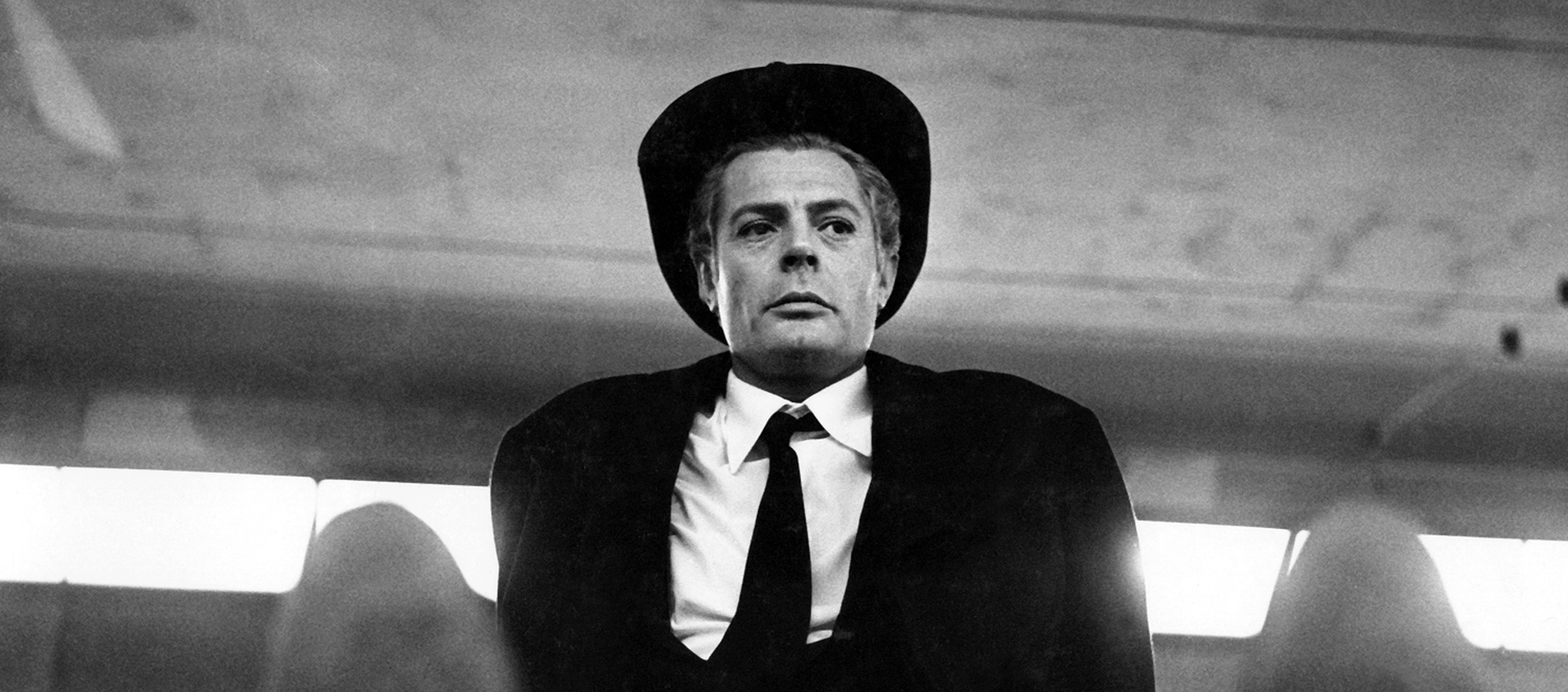 Black-and-white still of Guido (Marcello Mastroianni) sitting with legs outstretched, wearing a black suit, tie, and hat. A row of brightly lit windows is behind him.