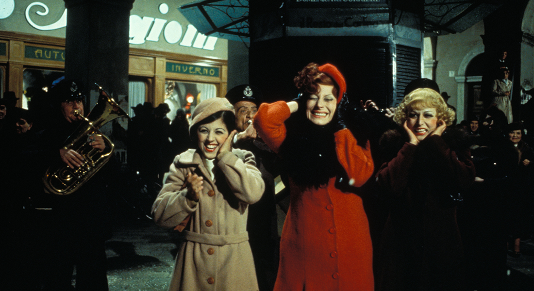 Still featuring Gradisca (Magali Noel), who is standing between two other women, all of whom are covering their ears as a band plays behind them. Gradisca wears a long red coat and matching hat.