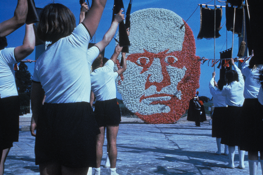 Still of school children in uniforms, holding up guns and flags as they face a huge papier-mache Mussolini sculpture.