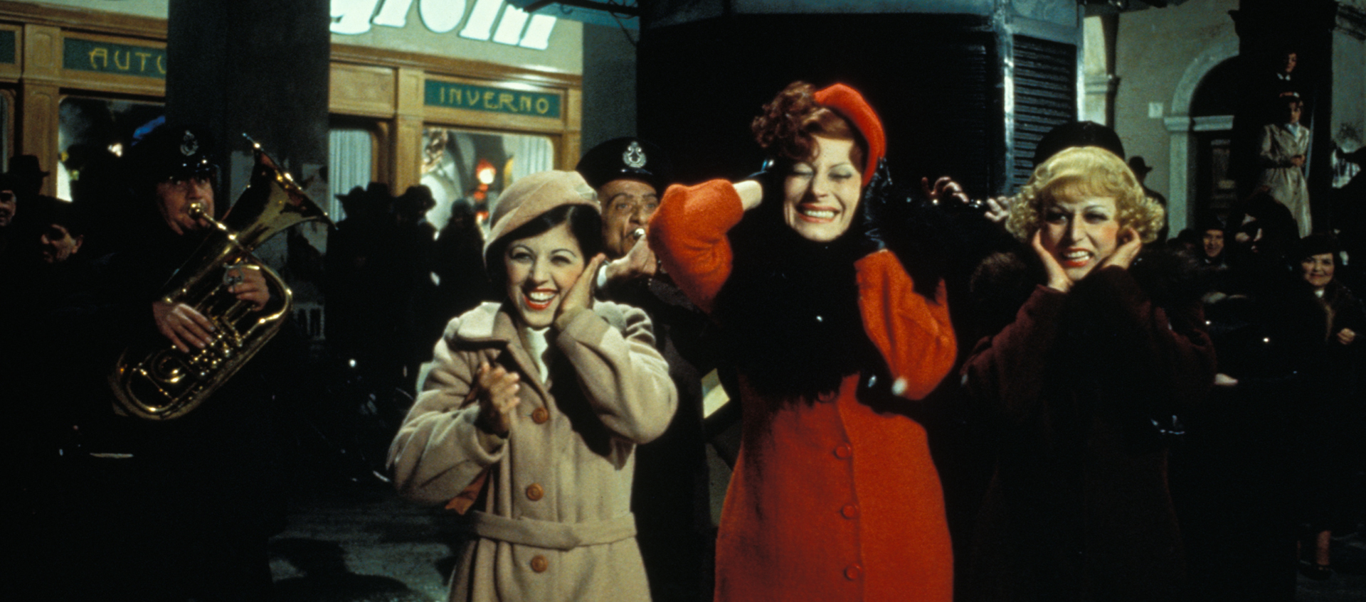 Still featuring Gradisca (Magali Noel), who is standing between two other women, all of whom are covering their ears as a band plays behind them. Gradisca wears a long red coat and matching hat.