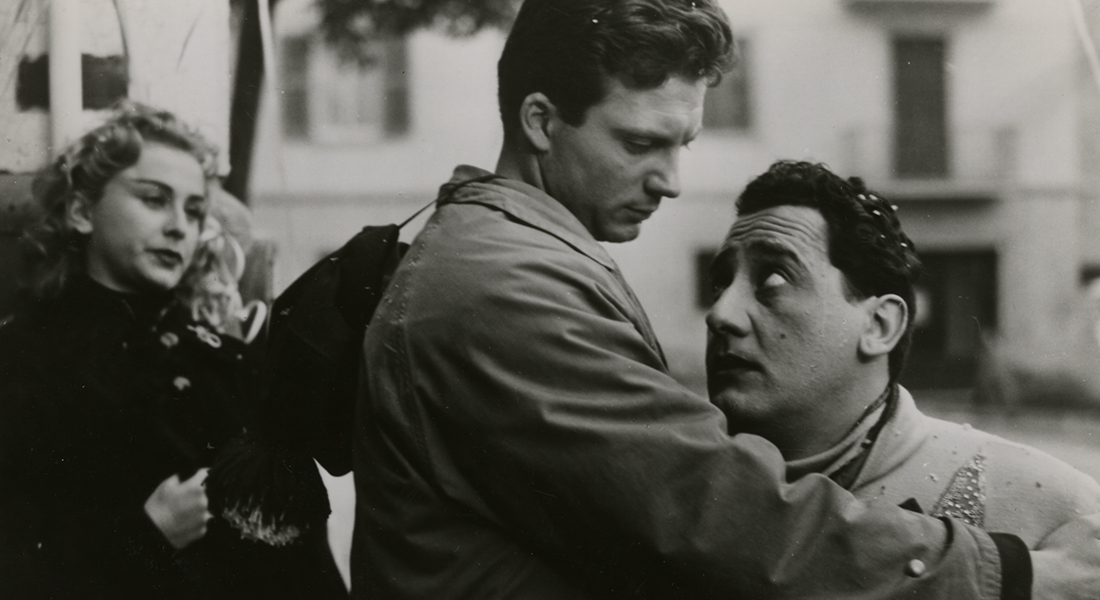 Black-and-white still from I vitelloni, featuring Fausto (Franco Fabrizi), Alberto (Alberto Sordi), and Gisella (Vira Silenti). Fausto (left) is looking down at Alberto, his hand on Alberto’s shoulder as if to console him. Gisella is standing in the background watching them.
