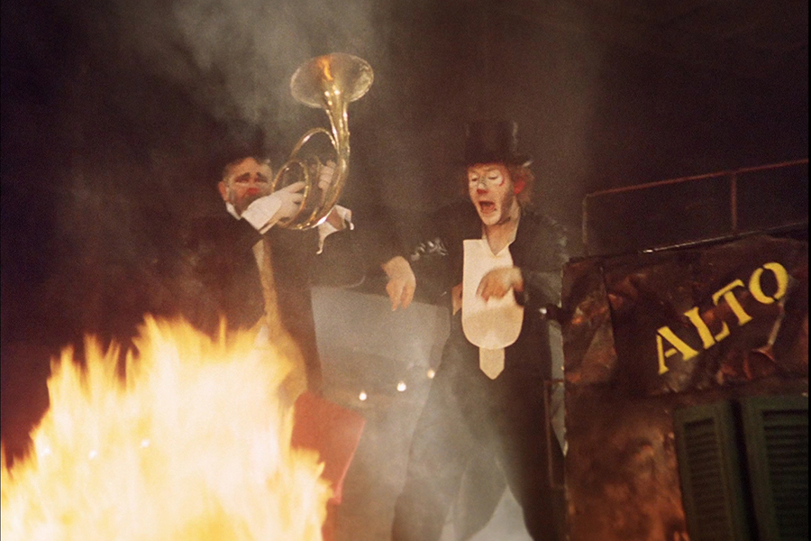 Still from I clowns featuring two people in clown makeup, black top hats, and tuxedos. The person on the left is playing a horn, and the person on the right has their mouth open and are pointing to the fire burning in front of them. The word “ALSO” in yellow is on a structure next to them.