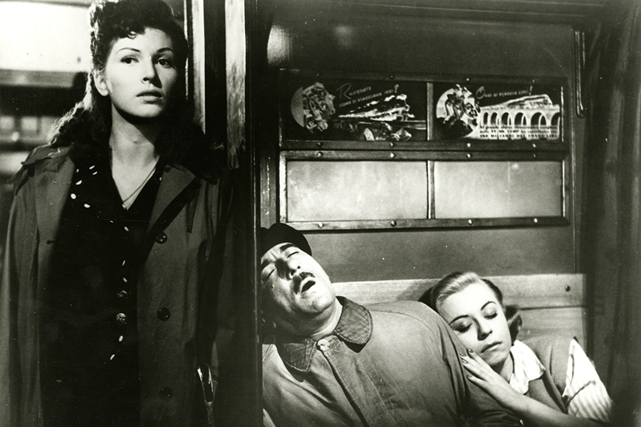 Black-and-white still featuring Lily (Carla Del Poggio) standing to the left of a seat where Checco (Peppino De Filippo) is sleeping with his head against the wall next to him. A woman to his right is sleeping against his shoulder.