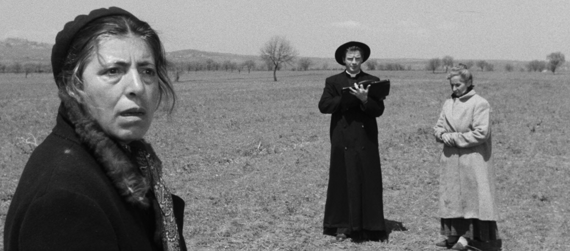 Black-and-white still featuring Maria (Stella Florina), Carlo (Richard Basehart), and actress Luccetta Muratori (from left to right). They are standing in a field; Maria is in the forefront with a worried look on her face. Farther in the distance is Carlo, who is dressed in a black priest robe and hat, and there is an older woman to his right; she is holding her hands together at her stomach and is looking down.