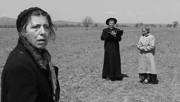 Black-and-white still featuring Maria (Stella Florina), Carlo (Richard Basehart), and actress Luccetta Muratori (from left to right). They are standing in a field; Maria is in the forefront with a worried look on her face. Farther in the distance is Carlo, who is dressed in a black priest robe and hat, and there is an older woman to his right; she is holding her hands together at her stomach and is looking down.