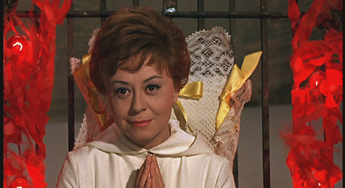Still of Giulietta (Giulietta Masina), who has short, light brown hair and is wearing a white blouse. Her hands are pressed together at her chest as if in prayer, and there are streams of red fire climbing up the metal-barred fence on either side of her and a doily with yellow ribbons against the fence behind her.