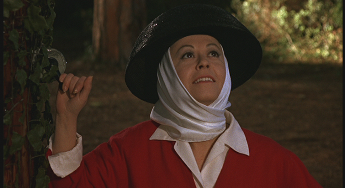 Still of Giulietta (Giulietta Masina), who is wearing a white scarf around her head, a back hat, and a red sweater over a white button-down shirt. She is standing next to a tree, one hand on a leaf, looking upwards and smiling.