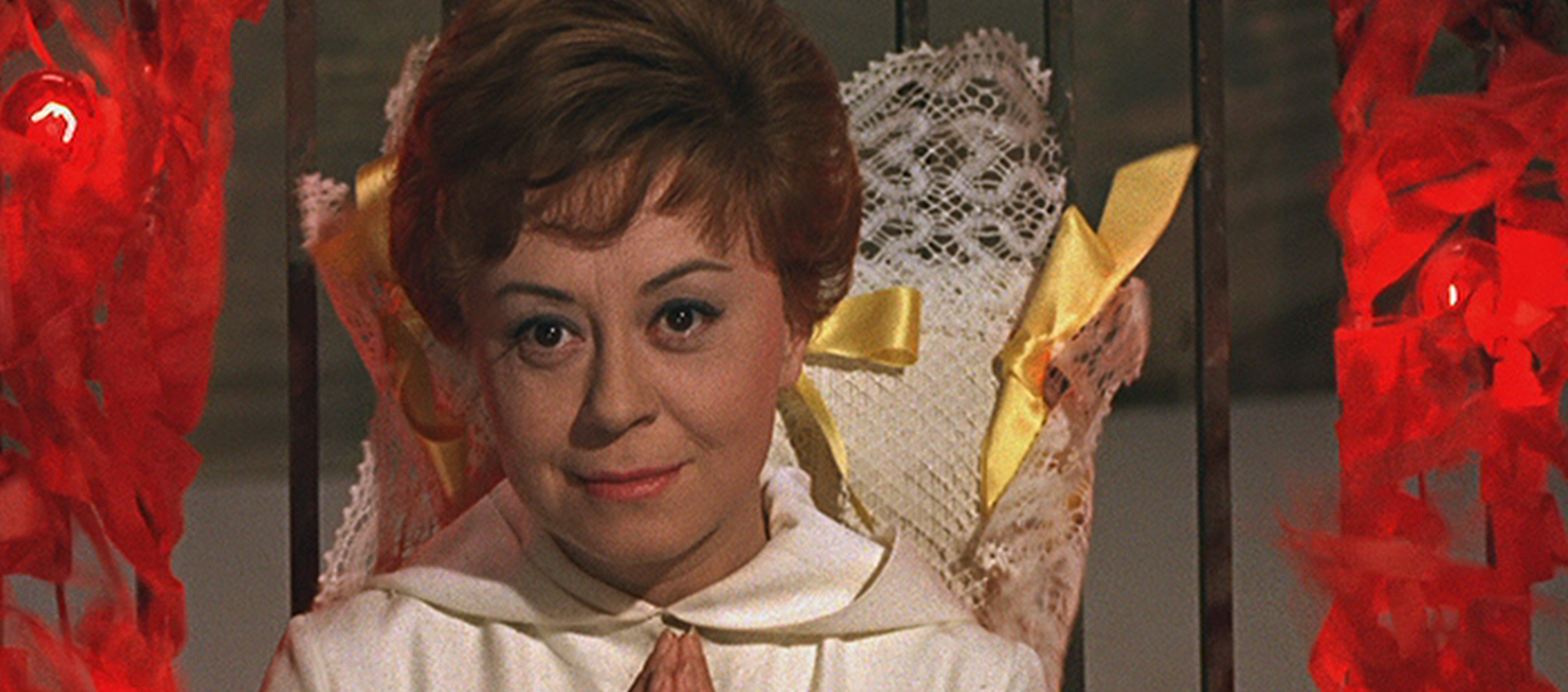 Still of Giulietta (Giulietta Masina), who has short, light brown hair and is wearing a white blouse. Her hands are pressed together at her chest as if in prayer, and there are streams of red fire climbing up the metal-barred fence on either side of her and a doily with yellow ribbons against the fence behind her.