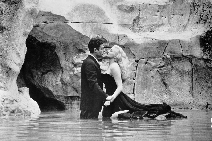 Black-and-white still of Marcello (Marcello Mastroianni) and Sylvia (Anita Ekberg) standing in the Trevi Fountain, facing each other. Marcello (left) has short dark hair, is wearing a dark suit, and is gripping Sylvia’s wrist. Sylvia (right) has long blonde hair and is wearing a dark ballgown that is floating in the water behind her.