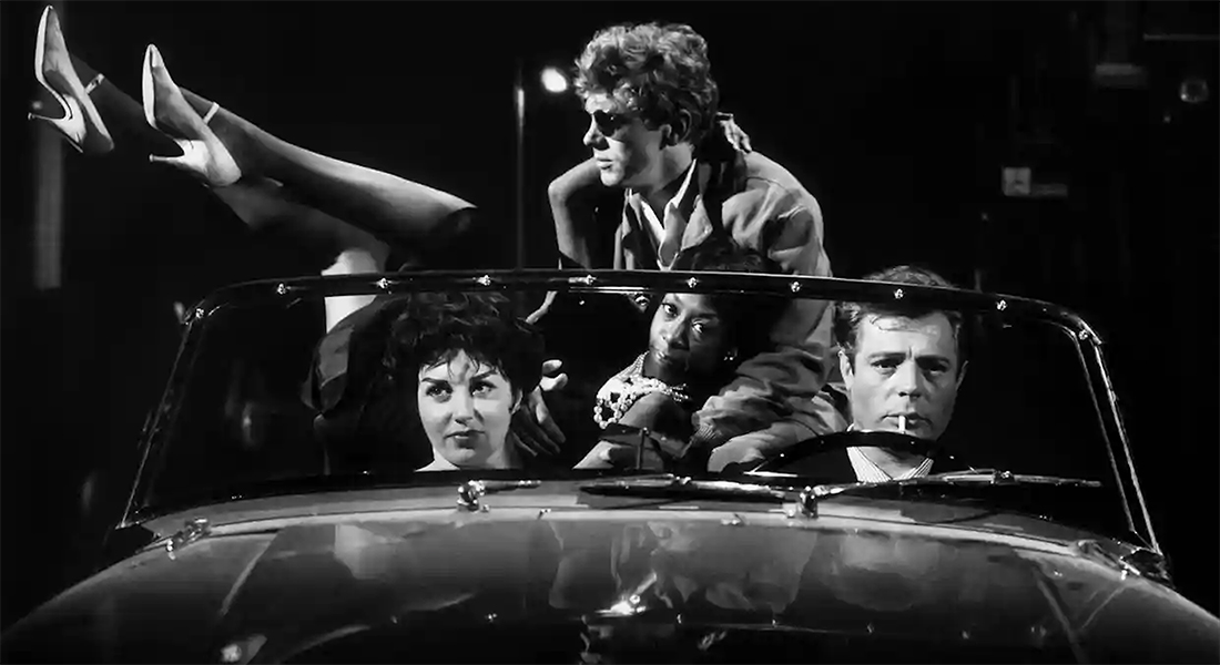 Black-and-white still of four people in a convertible; in the back, a couple sits on top of the car, one of whom is leaning against the other with their high heels in the air as they look into the camera.