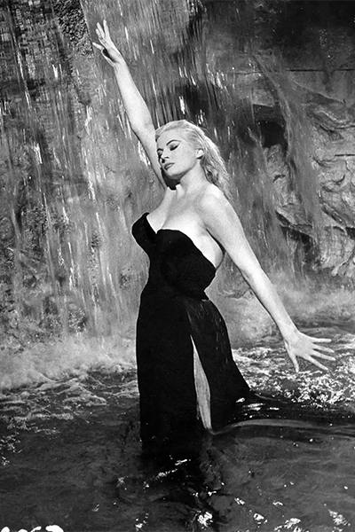 Black-and-white still of Sylvia (Anita Ekberg) standing in the Trevi Fountain wearing a dark ballgown. Her right arm is raised above her head, and her eyes are closed. A waterfall flows over a rock wall behind her.