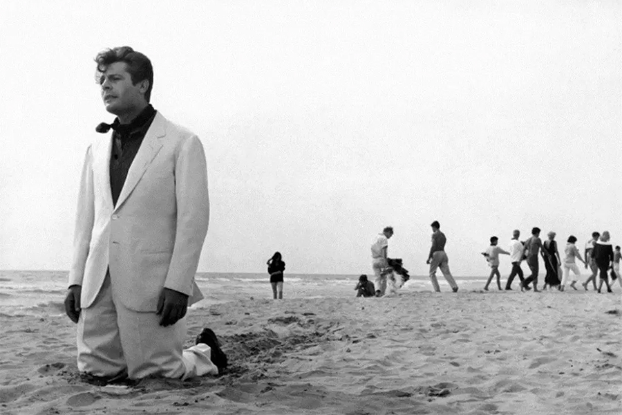 Black-and-white still of Marcello (Marcello Mastroianni) on his knees in the sand on the beach. He is wearing a white suit and looking into the distance. There is a group of people behind him. 