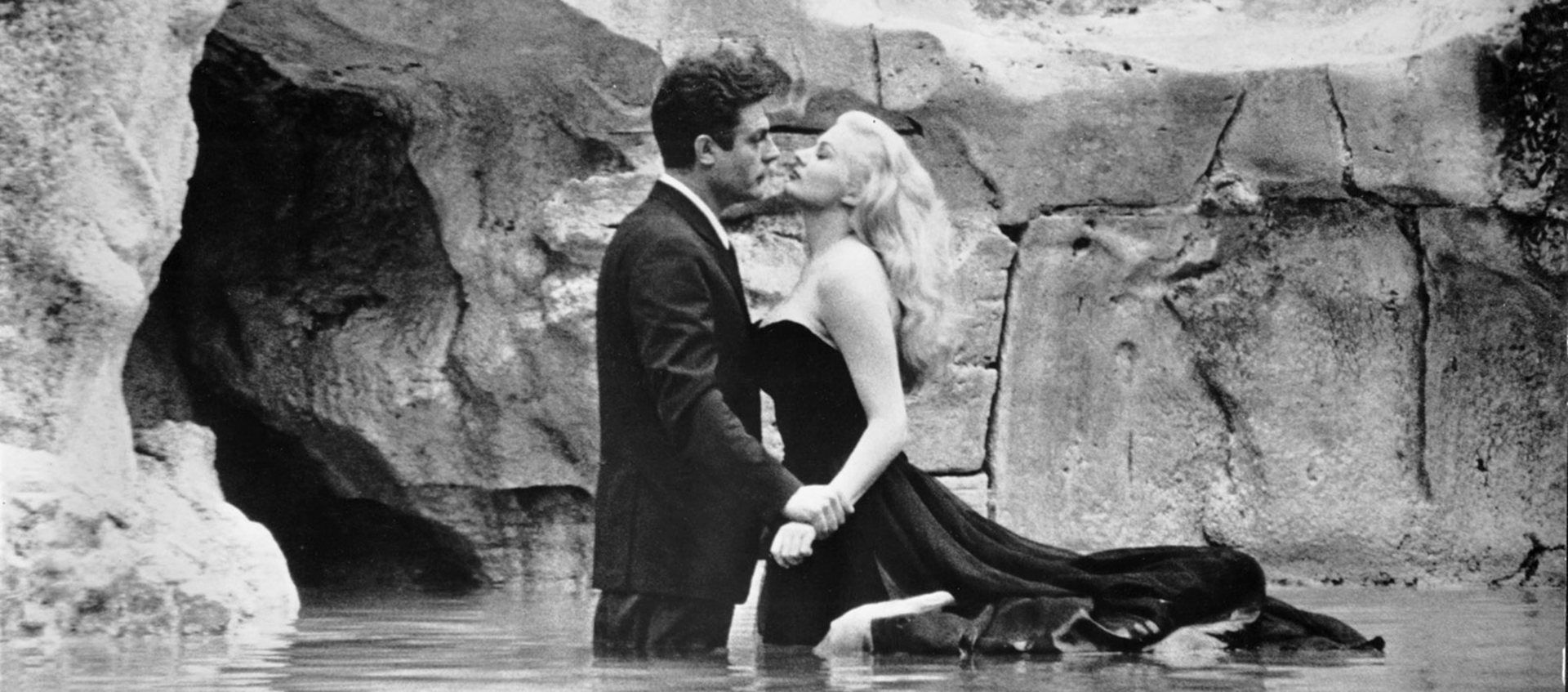 Black-and-white still of Marcello (Marcello Mastroianni) and Sylvia (Anita Ekberg) standing in the Trevi Fountain, facing each other. Marcello (left) has short dark hair, is wearing a dark suit, and is gripping Sylvia’s wrist. Sylvia (right) has long blonde hair and is wearing a dark ballgown that is floating in the water behind her.