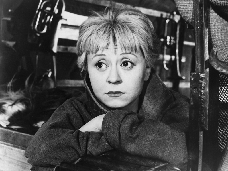 Black-and-white still of Gelsomina (Giulietta Masina), who is resting her crossed arms on the back door of the vehicle she is in, looking into the distance with widened eyes and raised eyebrows as if concerned.