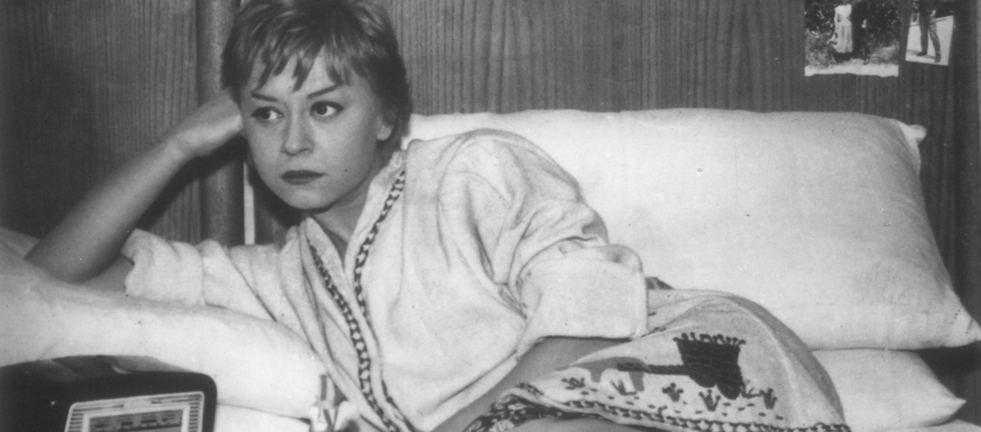 Black-and-white still of Cabiria (Giulietta Masina) lying on a bed with her head resting against her fist and her elbow on the pillow. There is a radio on the bed next to her and photographs on the headboard.